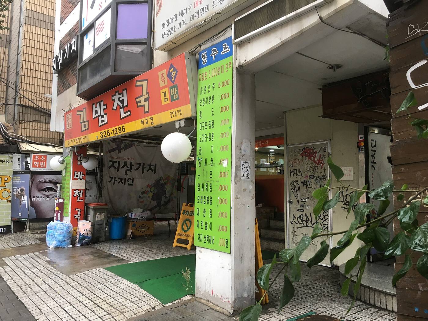 The street front of a Kimbap Cheonguk branch in Seoul, orange sign with the chain name and a small 24-hour sticker, next to a green price board for a neighbouring bar.