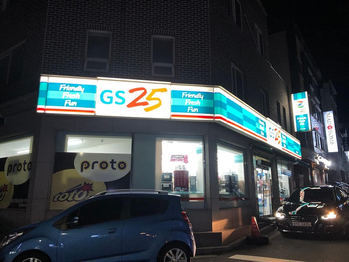 A GS25 convenience store on a street corner in South Korea at night, its blue-and-white sign lit, with a car parked in front.