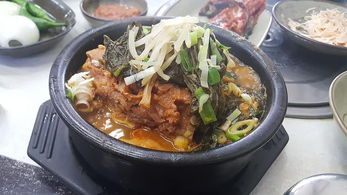A black stone bowl of ppyeodagwi haejangguk, pork-bone hangover soup, topped with bean sprouts, dried cabbage leaves and spring onion, with side dishes on the table.