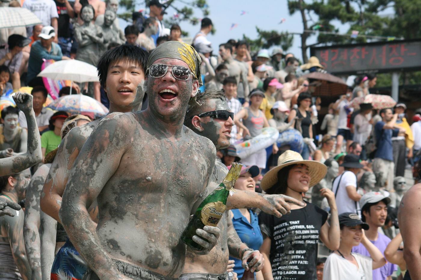 Laughing, mud-smeared festival-goers, one holding a champagne bottle, in front of a crowd on the steps at the Boryeong Mud Festival, Daecheon Beach.