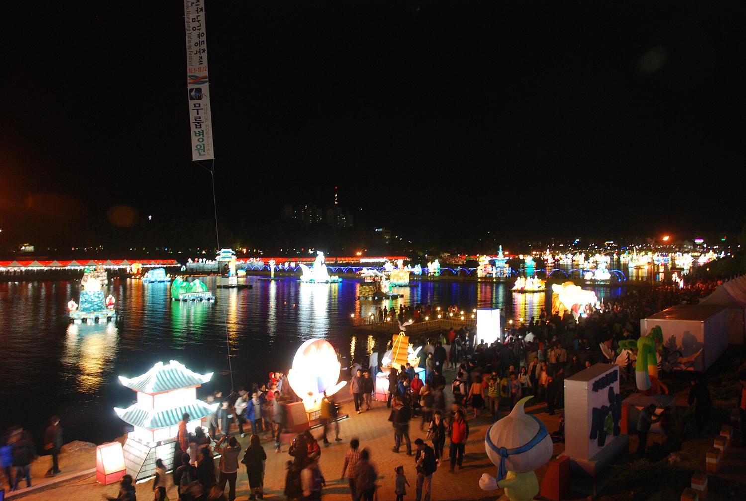Illuminated lantern floats on the Nam River at night during the Jinju Namgang Yudeung Festival, with a lit lantern bridge across the water, crowds on the bank and a festival banner overhead.