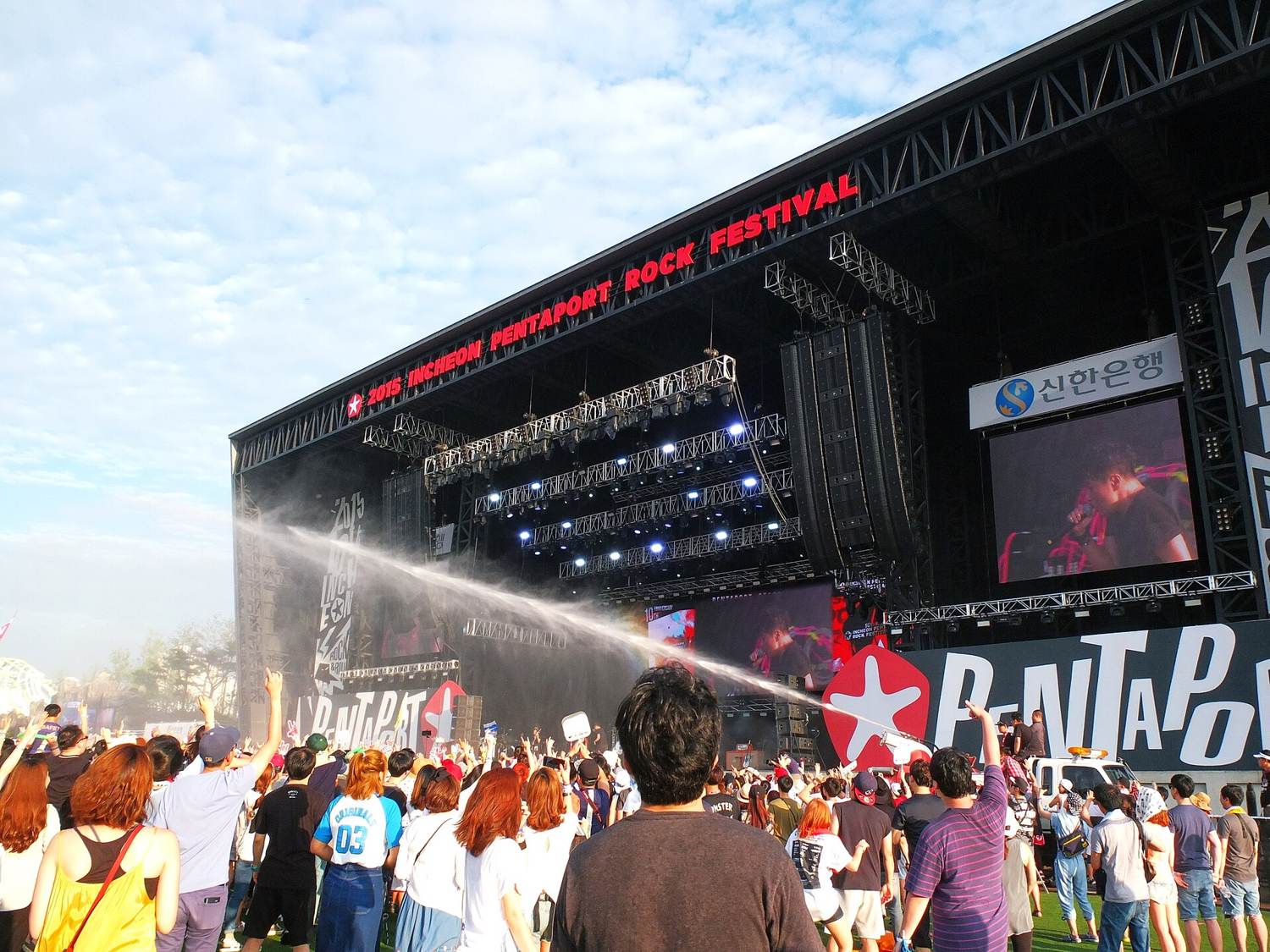 The main stage of the 2015 Incheon Pentaport Rock Festival at Songdo, a crowd with raised arms in front of the stage sign and a water cannon spraying the audience.