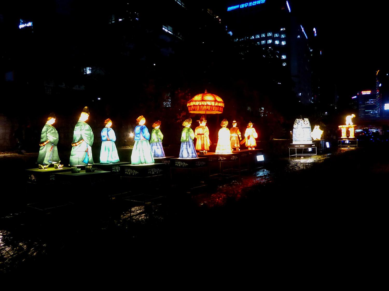 A procession of illuminated lantern figures in Joseon court dress standing in Cheonggyecheon stream at night during the Seoul Lantern Festival, office towers lit behind.
