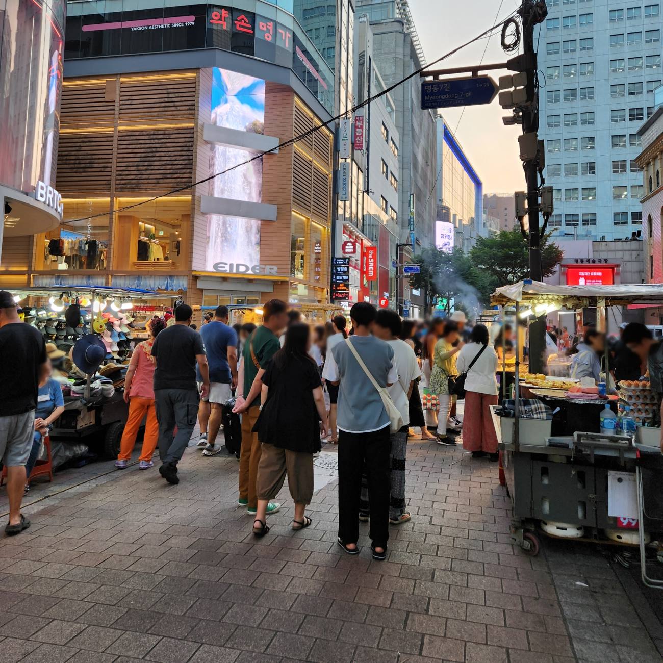 A crowded evening street in Myeongdong, Seoul, with street-food carts, a hat stall and shoppers walking between them under shop signs.