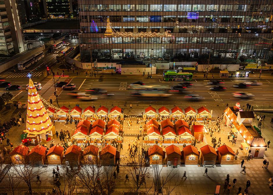 Aerial night view of the 2024 Gwanghwamun Market: rows of red-roofed wooden huts lit with string lights on Gwanghwamun Square, a tall lit Christmas tree at one corner, traffic blurred on Sejong-daero beside it.