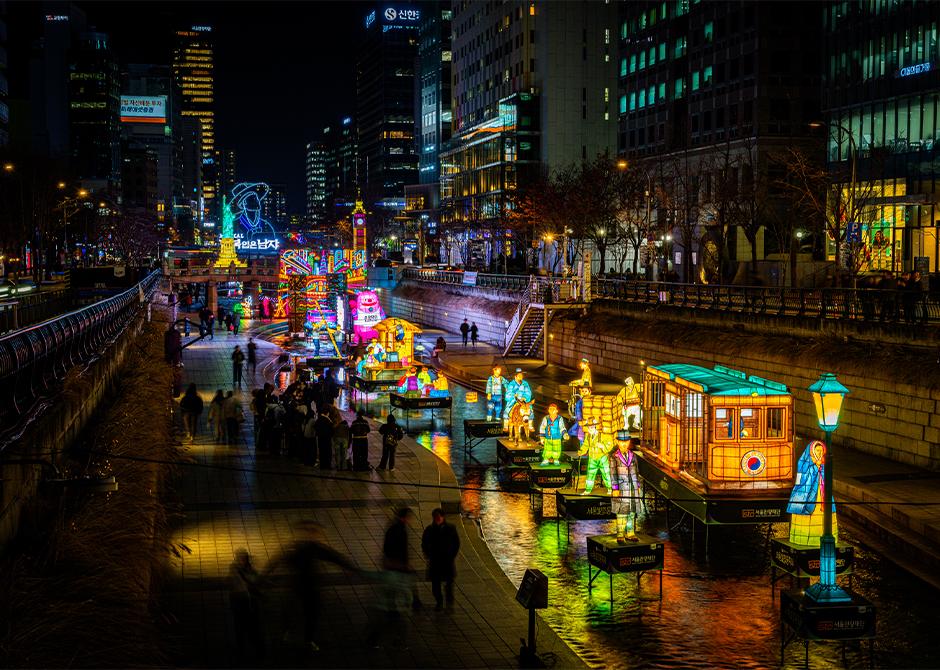 The 2025 Seoul Lantern Festival on Cheonggyecheon at night: illuminated lantern figures and a lantern tram on the water, crowds along the lower walkway, office towers lit behind.