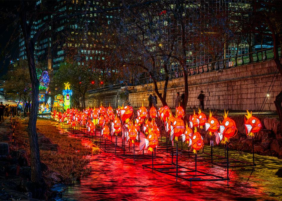 A long row of orange Magikarp Pokémon lanterns in Cheonggyecheon stream at night, lit red, with the stone embankment and bare trees behind.