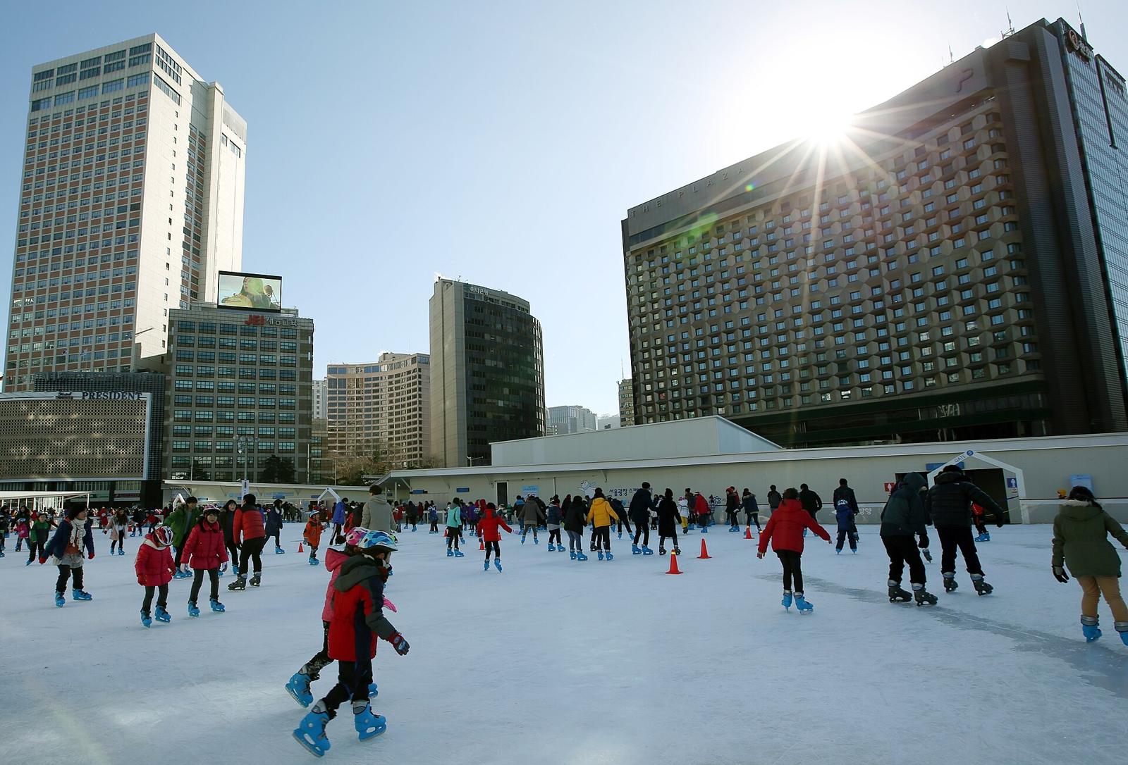 Skaters on the outdoor Seoul Plaza ice rink in front of City Hall on a sunny winter day, children in helmets in the foreground, the Plaza Hotel and office towers behind.