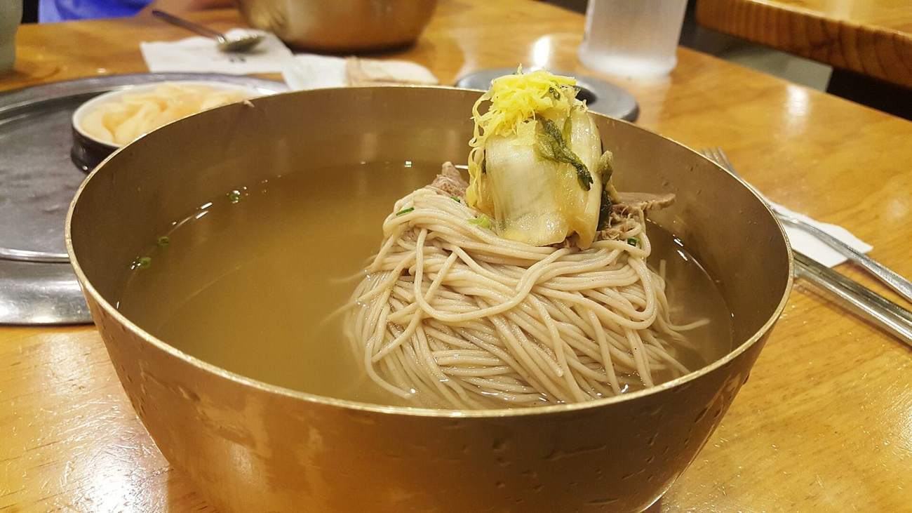 A brass bowl of Pyongyang-style naengmyeon: buckwheat noodles in clear cold broth with sliced beef and a piece of kimchi under shredded egg.