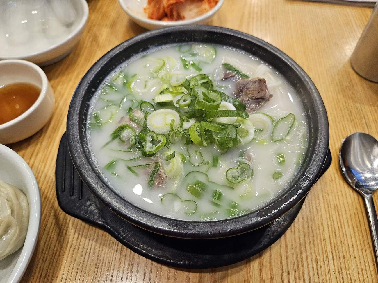 A black stone bowl of milky-white seolleongtang with beef, thin noodles and sliced spring onion on top, cabbage kimchi and dumplings beside it.