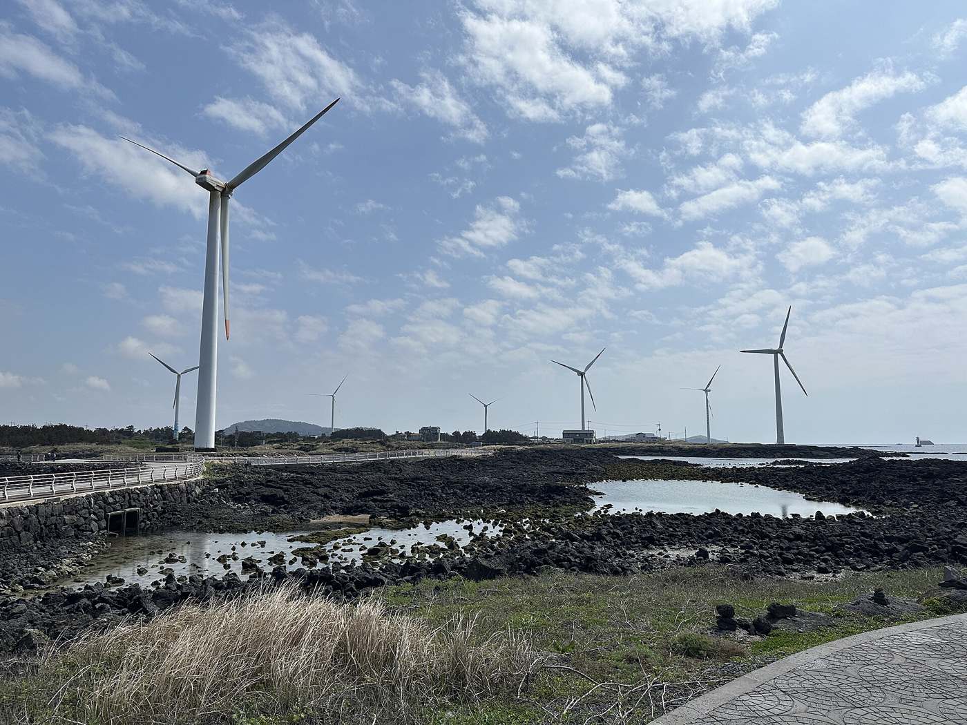 Wind turbines along the lava-rock shore at Sinchang on Jeju's west coast, with the coastal road behind its white guardrail on the left and a paved promenade in the foreground.