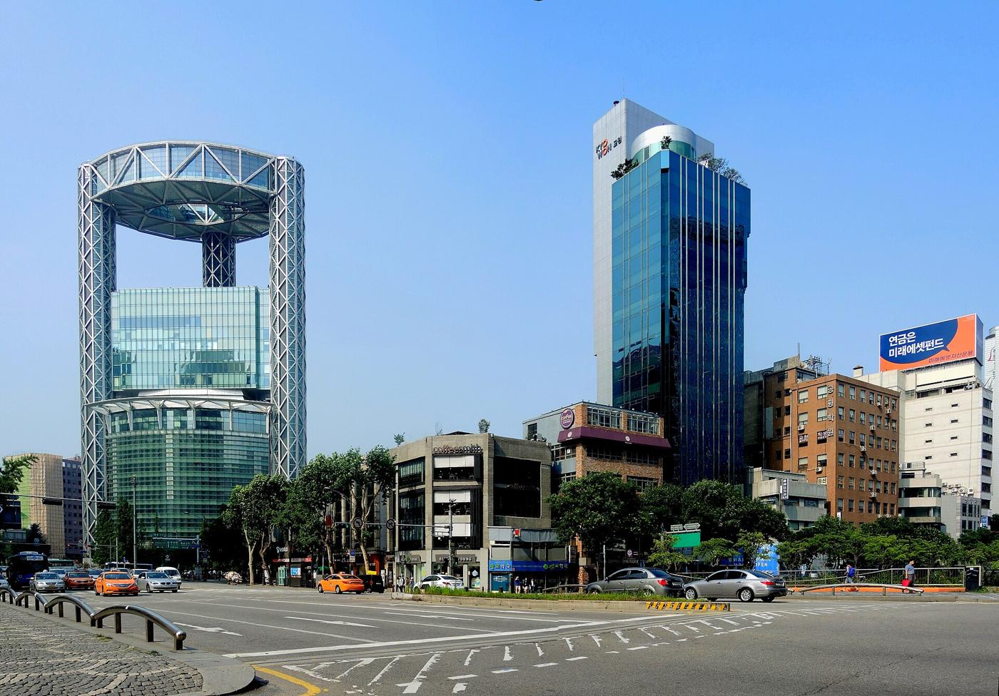 Jongno Tower in central Seoul, a glass tower inside a steel frame with a ring-shaped top, beside a blue glass office building across a wide junction.