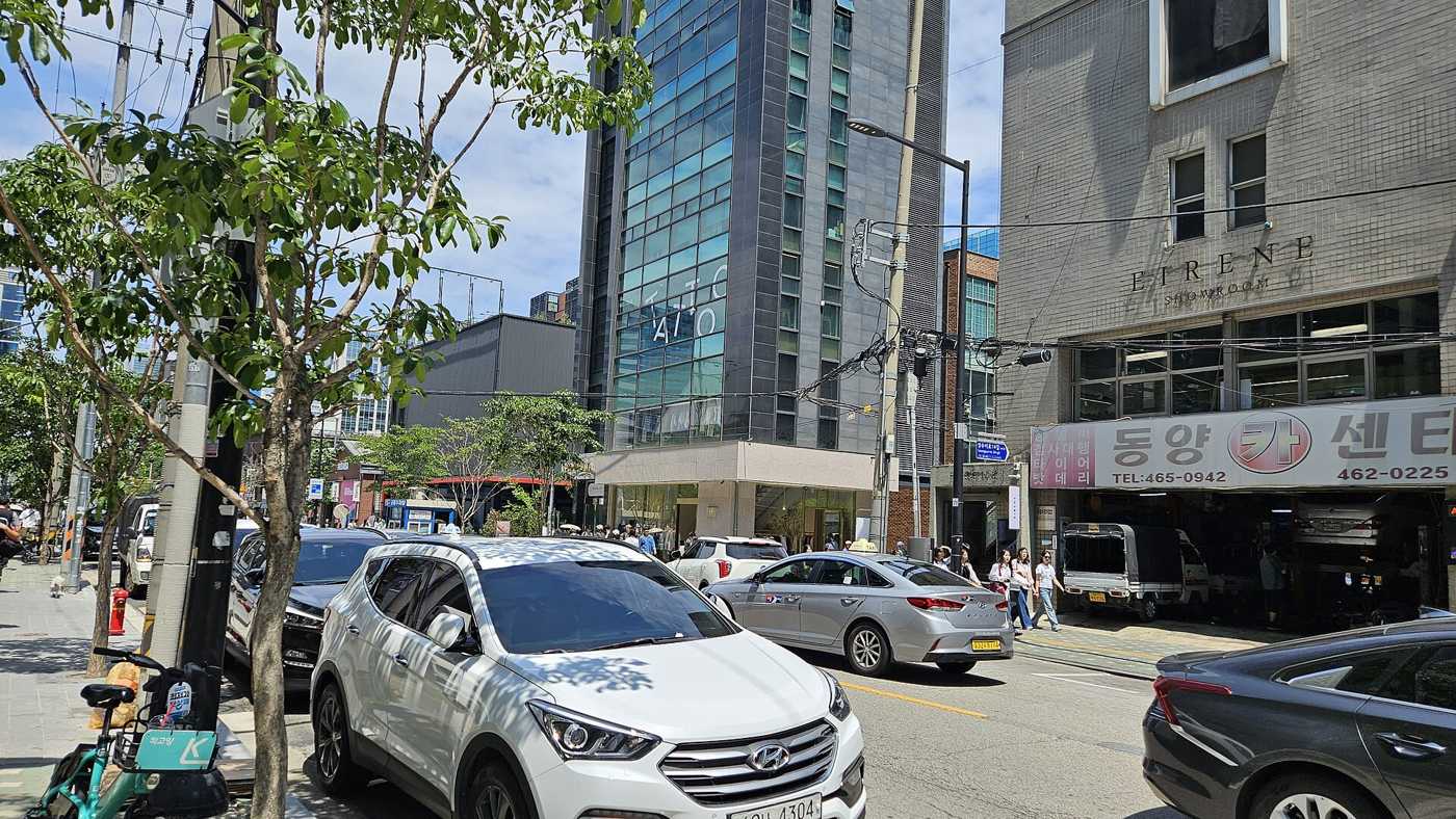A street in Seongsu-dong, Seoul, in May 2026: a glass office tower, a car-repair garage and a showroom building, with cars parked along the kerb.