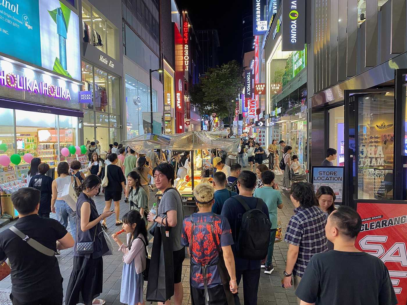 Myeongdong's night market street in Seoul in August 2024, a lit food cart and shoppers filling the road between the shopfronts.
