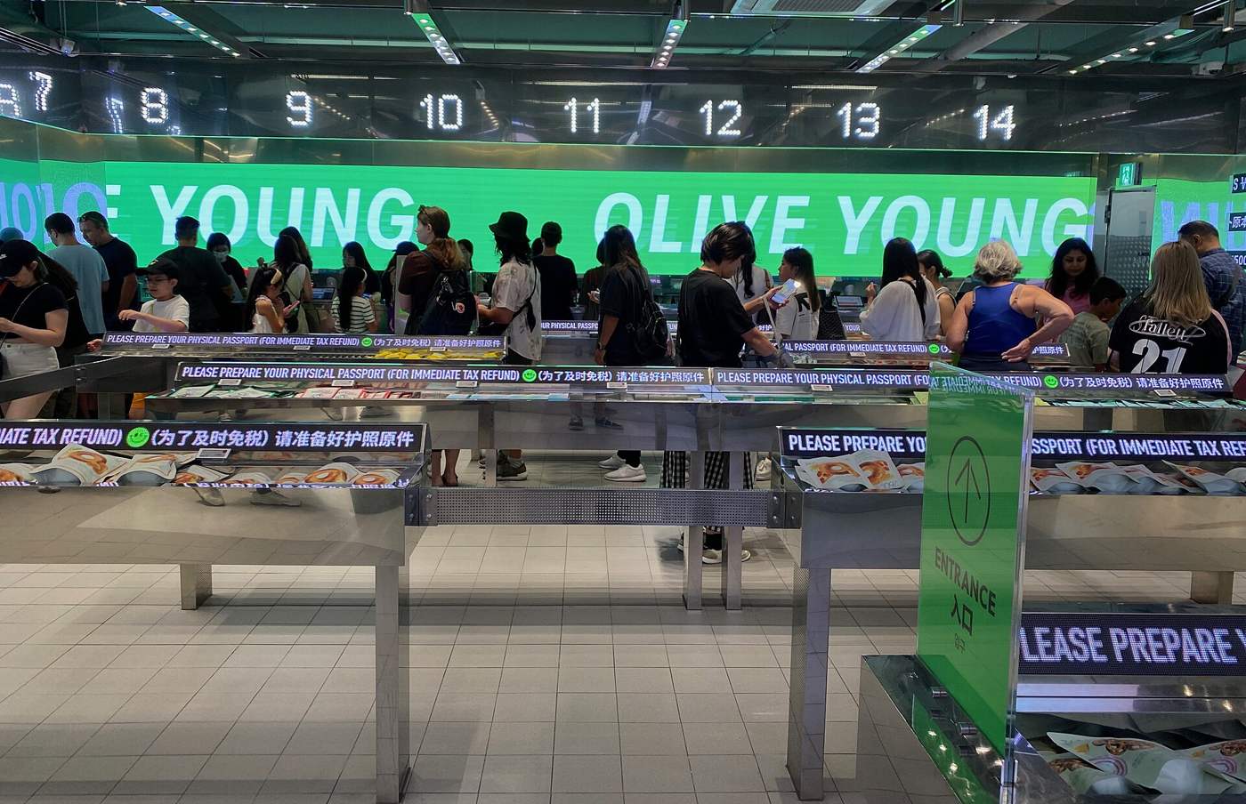 Checkout counters inside Olive Young's Myeongdong store in August 2024, with tax-refund passport signs and a queue of shoppers under a green OLIVE YOUNG screen.