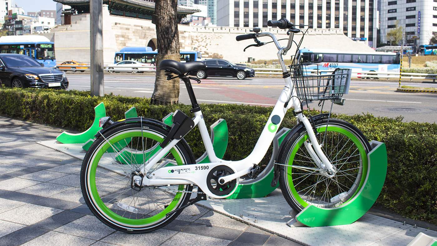 A QR-type "new Ttareungi" docked at a green rack on a Seoul pavement, with city traffic behind it.