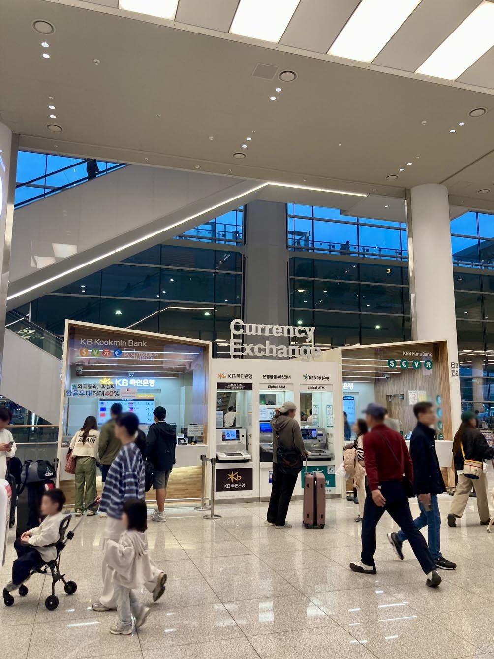 Two currency exchange booths in Incheon Airport Terminal 2, KB Kookmin Bank on the left and KEB Hana Bank on the right, with three Global ATMs between them and a Korean sign on the KB booth advertising a preferential exchange rate of up to 40 percent.