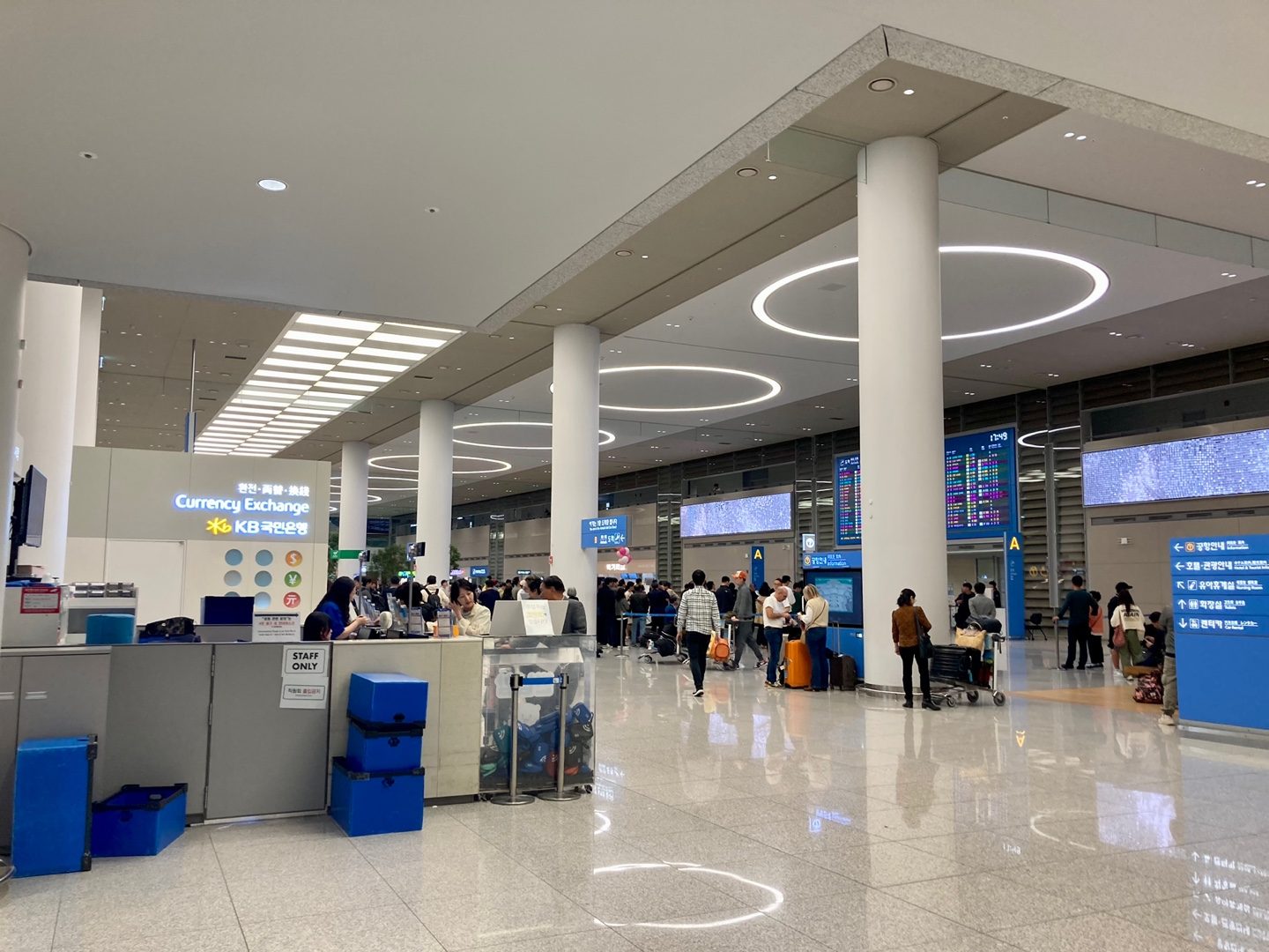 The arrivals hall of Incheon Airport Terminal 2 in October 2024, with a KB Kookmin Bank counter under a lit 'Currency Exchange' sign on the left and travellers with suitcases walking past the flight boards.