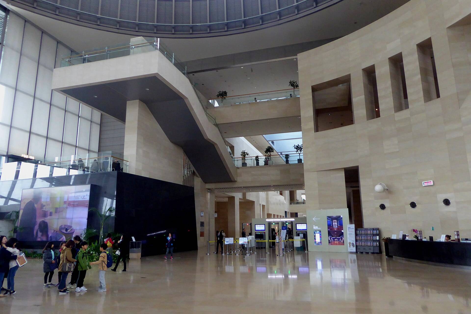 The entrance hall of the National Museum of Korea, a tall stone atrium with a staircase and balconies, security gates and an information desk, and a school group waiting on the polished floor.