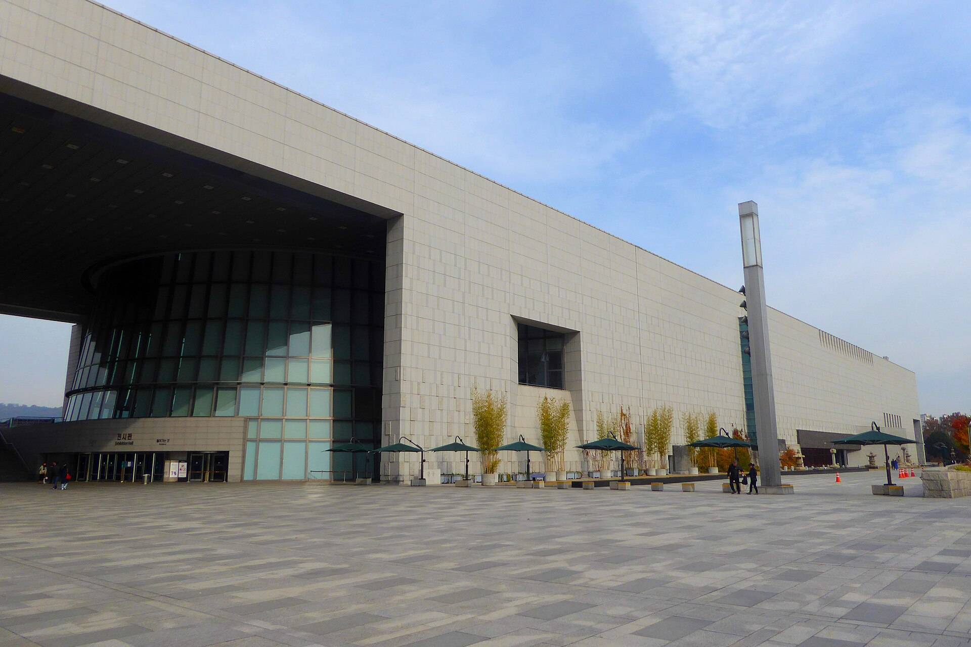 The long white stone façade of the National Museum of Korea in Yongsan, Seoul, with the open central gateway on the left, a wide paved plaza lined with green parasols and a few visitors.