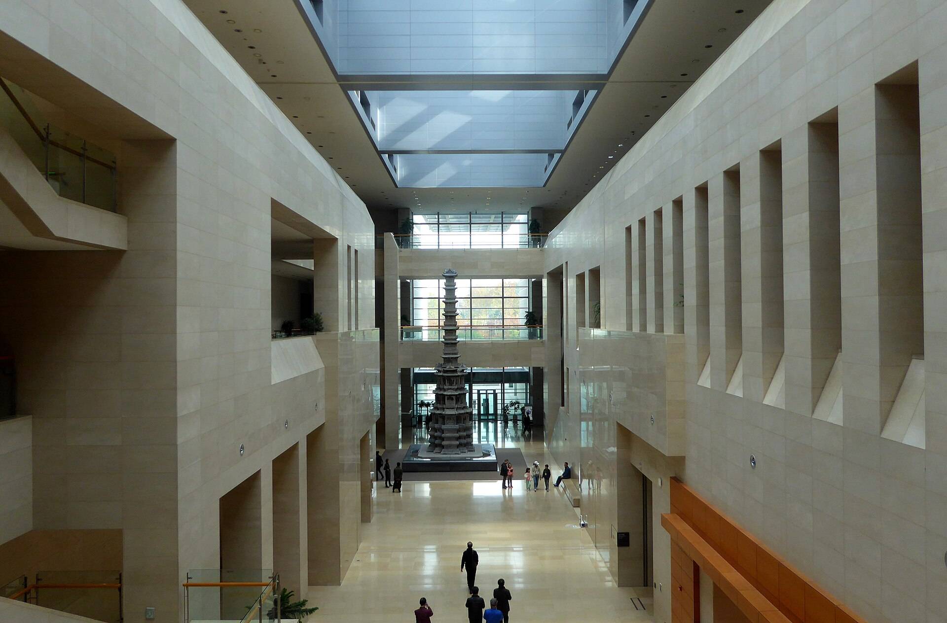The long central hall of the National Museum of Korea under a glass ceiling, with the grey ten-storey stone pagoda from Gyeongcheonsa Temple standing at the far end and visitors walking towards it.