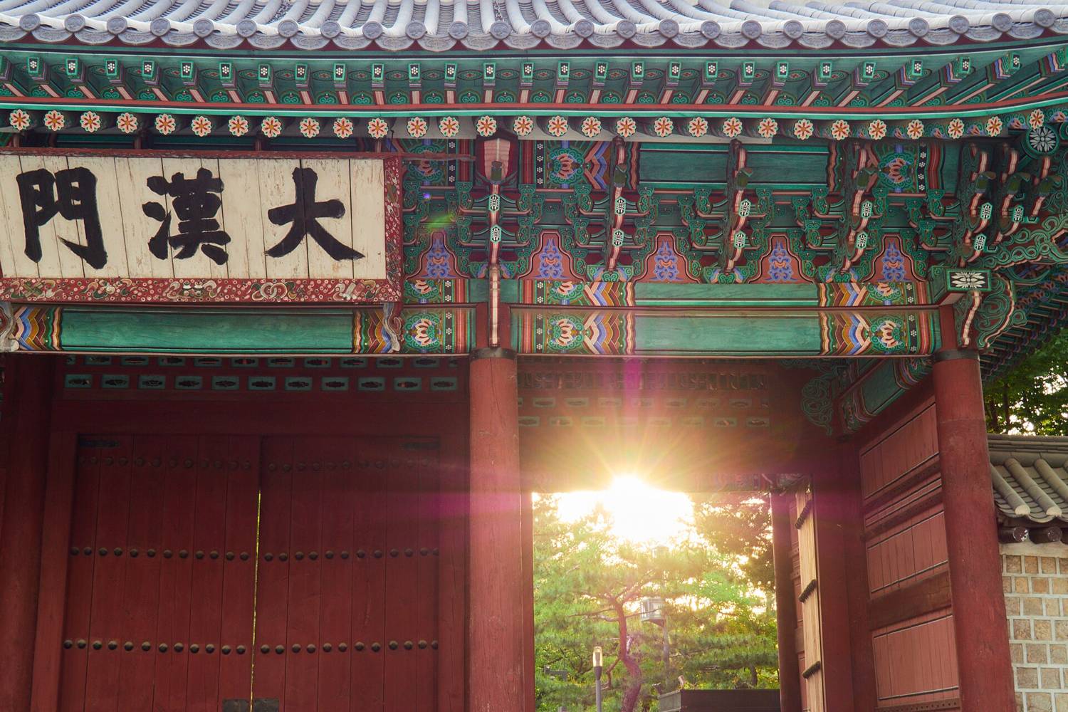 The main gate of Deoksugung, Daehanmun, with a signboard reading 大漢門 in black characters, painted dancheong eaves and the red gate doors, with sunlight coming through the open gateway.