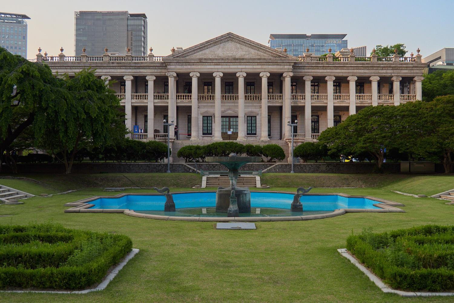 The Seokjojeon stone hall at Deoksugung, a neoclassical building with Ionic columns, with a formal garden and a fountain in front and modern office towers behind.