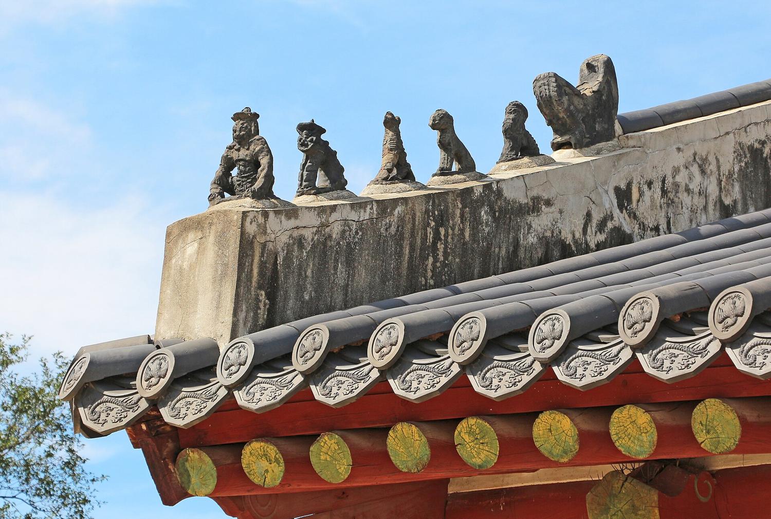 Clay guardian figures lined up along the roof ridge of Jongmyo's entrance gate, above grey roof tiles and red-painted rafters, against a blue sky.
