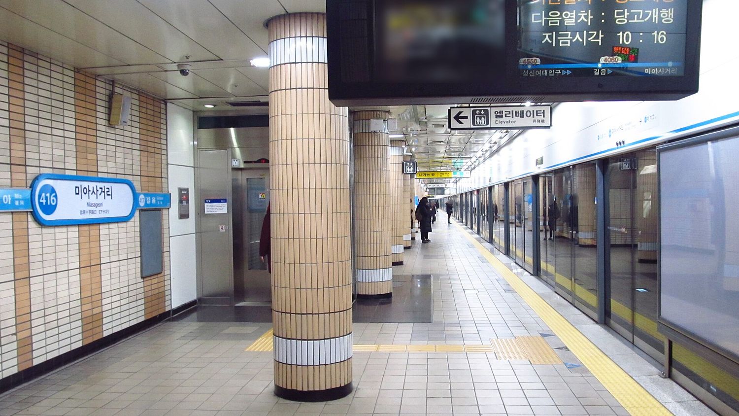 A Seoul subway platform at Miasageori station with a stainless-steel elevator on the left, a hanging sign reading 엘리베이터 Elevator with an arrow, and platform screen doors on the right.