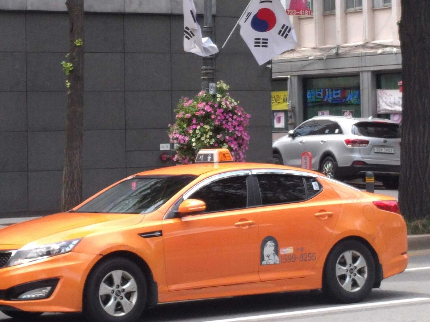 An orange Seoul taxi under Korean flags on a city street, with its roof sign reading 서울 (Seoul).