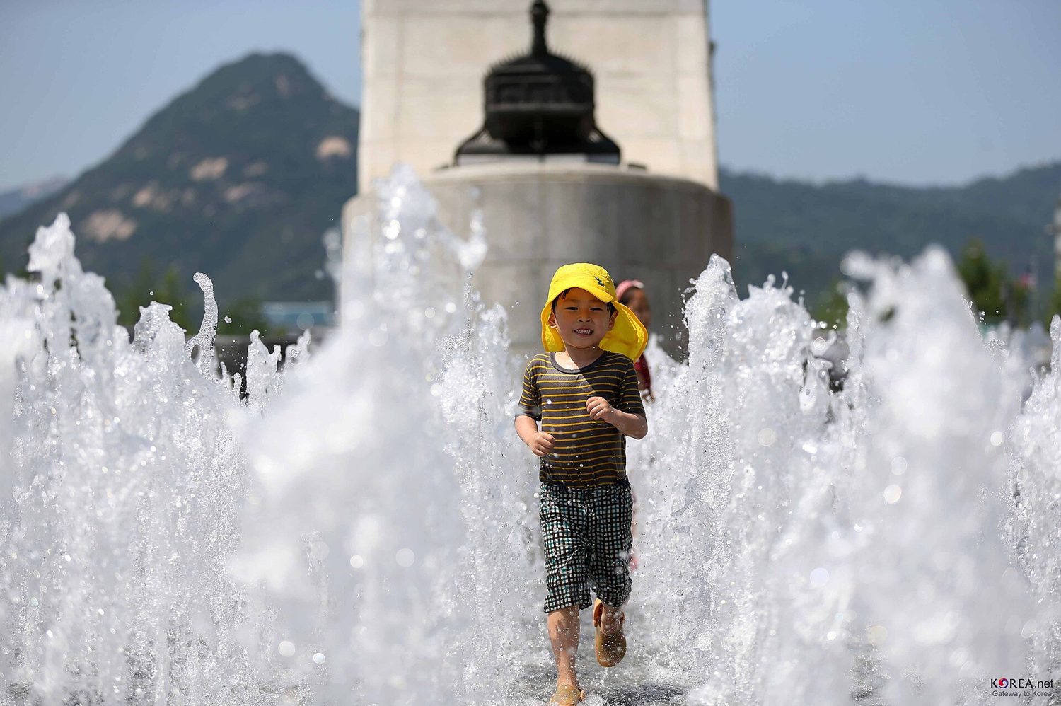 A small boy in a yellow sun hat running through the fountain jets on Gwanghwamun Square in Seoul, with the base of a statue and Bugaksan mountain behind him.