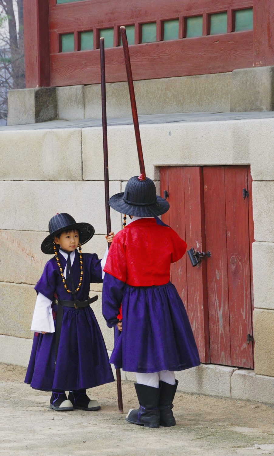 Two small children dressed as Joseon palace guards in purple and red robes and black hats, holding tall staffs, standing by a red door at Gyeongbokgung Palace.