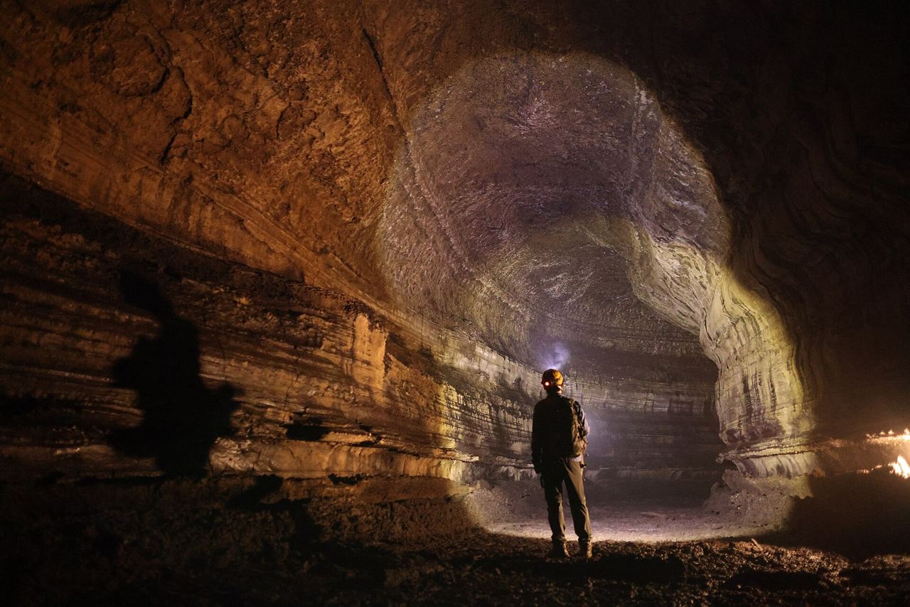 A person wearing a headlamp standing in Manjanggul's huge arched lava tube passage, with horizontal lava shelves along the walls.