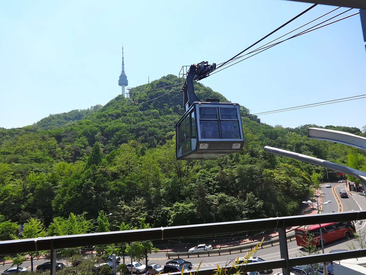 A grey Namsan Cable Car cabin leaving the lower station, with forested Namsan and N Seoul Tower behind it.