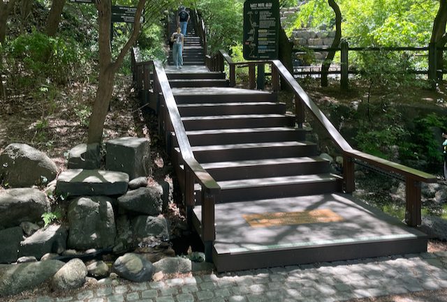 The foot of the Northern Forest Trail on Namsan, a wooden deck staircase rising through trees beside a pile of stones.
