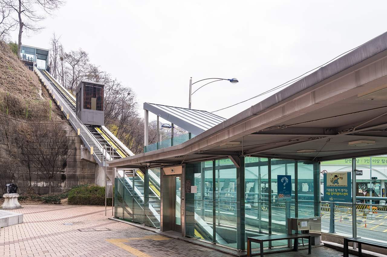 The lower station of the Namsan Oreumi incline elevator, a glass-walled waiting area with a small cabin on the incline rail climbing the slope behind.