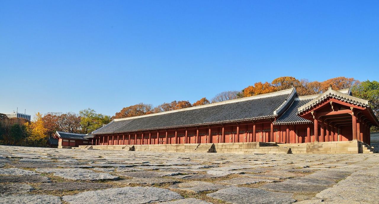 Jongmyo's main shrine hall, Jeongjeon, a long row of red columns under a single roof above a wide stone courtyard, with autumn trees behind.