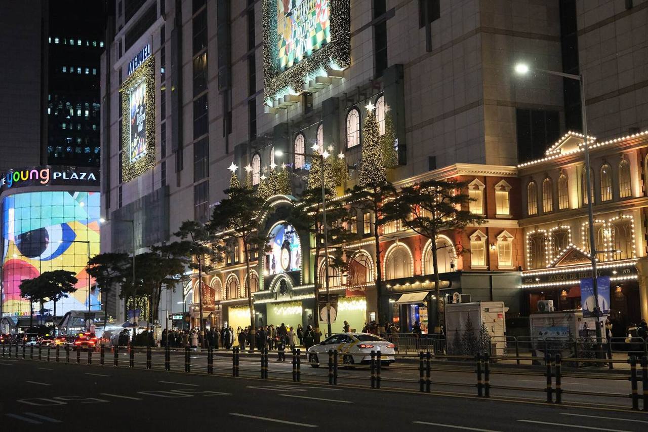 Lotte's flagship building in Myeongdong at night, lit with the AVENUEL sign and Christmas lights, taxis and pedestrians on the street in front.