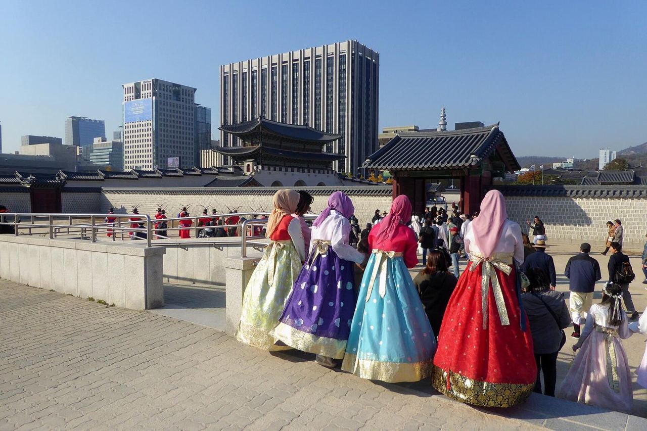 Four women wearing headscarves and hanbok, seen from behind, watching the guard ceremony inside Gyeongbokgung, with Gwanghwamun gate and office towers behind.