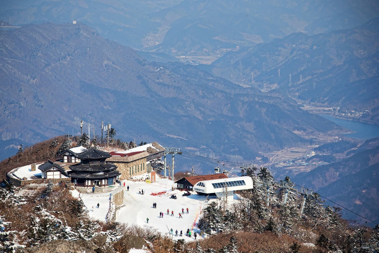 The summit station at Seolcheonbong on Deogyusan, seen from a higher peak: a snow-covered ridge with a traditional Korean tiled pavilion, a stone lodge, the arched glass top station of the Deogyusan gondola with cabins on the cable, and skiers walking on the packed snow, with hazy blue ranges and a reservoir far below.