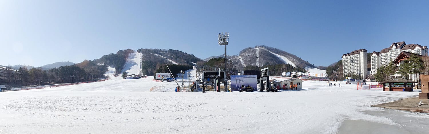 A wide panorama of Yongpyong Resort's ski base on a clear day: groomed white slopes rising into forested ridges, a chairlift line, Korean-language sponsor banners at the base, and the resort's condominium towers on the right.