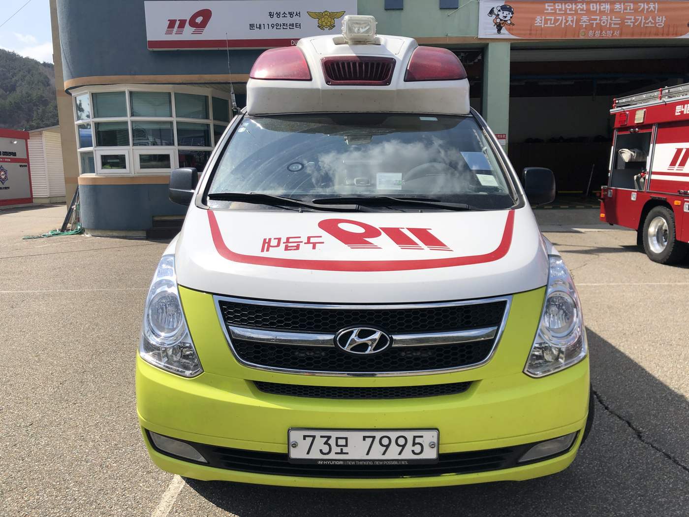 A Hyundai 119 ambulance in fire-service white and lime green, parked in front of the Dunnae 119 Safety Center with a fire engine behind it.