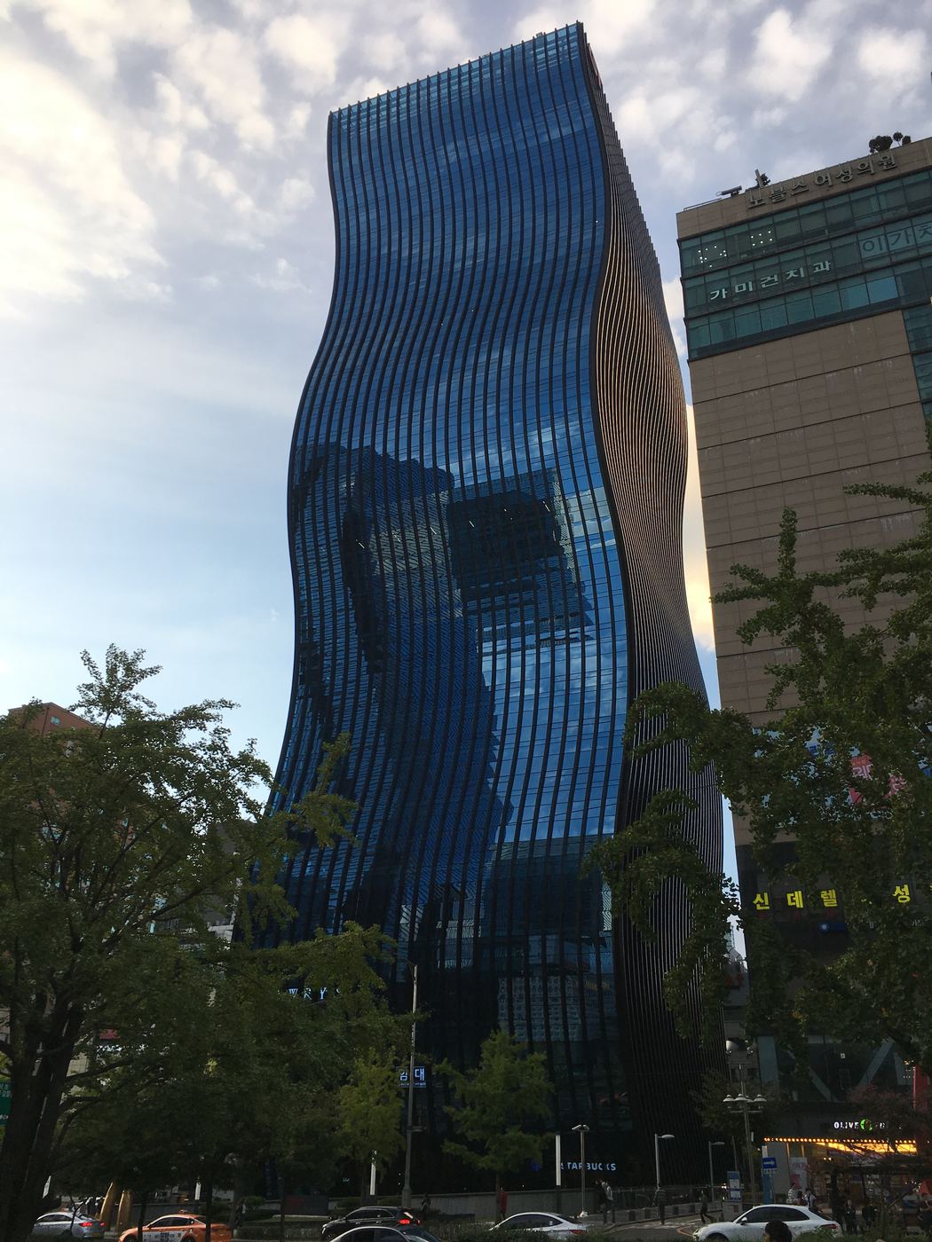 GT Tower, a tall curved blue-glass skyscraper beside Gangnam Station in Seoul, with a neighbouring building lettered with Korean clinic signs including 노블스여성의원 and 가미건치과.