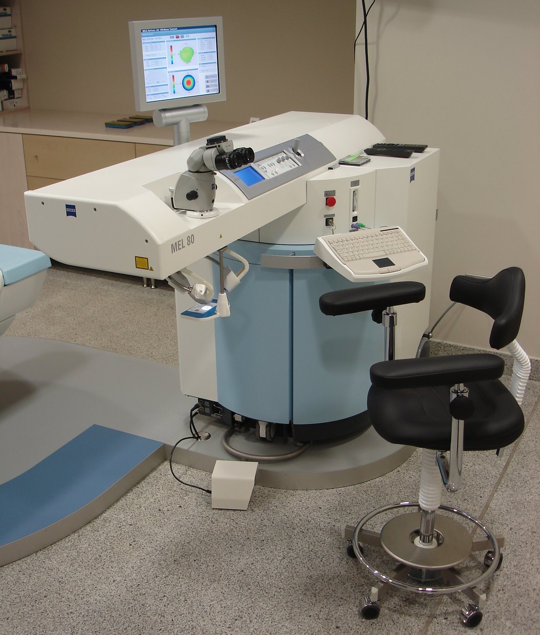 A Zeiss MEL 80 excimer laser in a treatment room: a white and blue console with a surgical microscope, a monitor showing corneal maps, and a surgeon's chair.