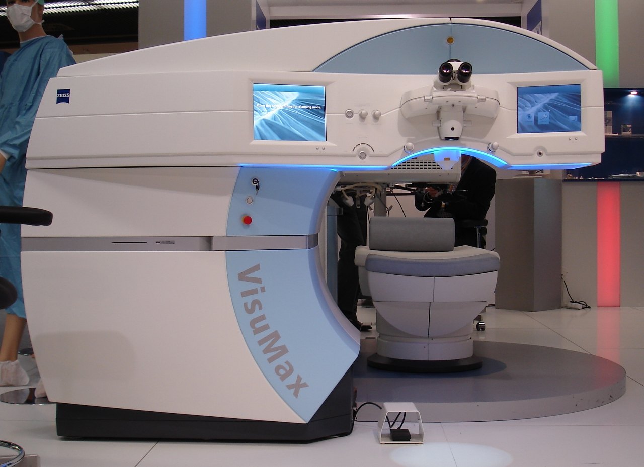 A Zeiss VisuMax femtosecond laser on display: a white console with two monitors, a surgical microscope above a patient bed, and the word VisuMax printed on the side.