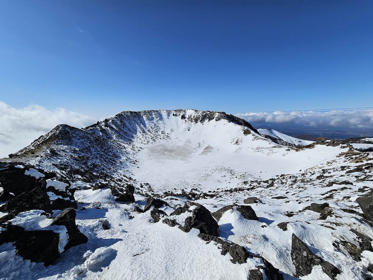 Baengnokdam, Hallasan's summit crater, filled with snow under a deep blue sky in January, with dark volcanic rocks on the rim and clouds below.