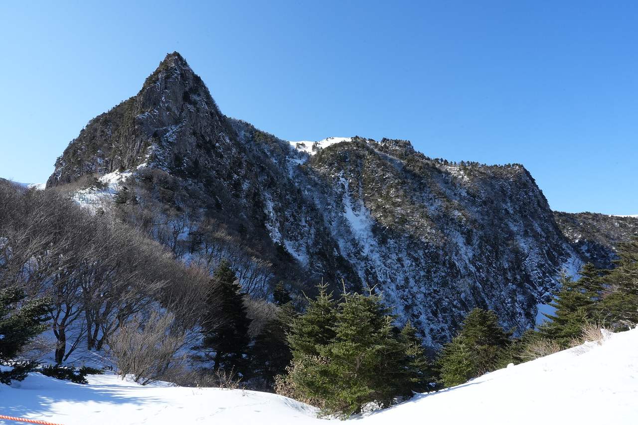 The triangular rock peak of Samgakbong on Hallasan under a clear blue sky, with snow on its slopes and a stand of conifers below.