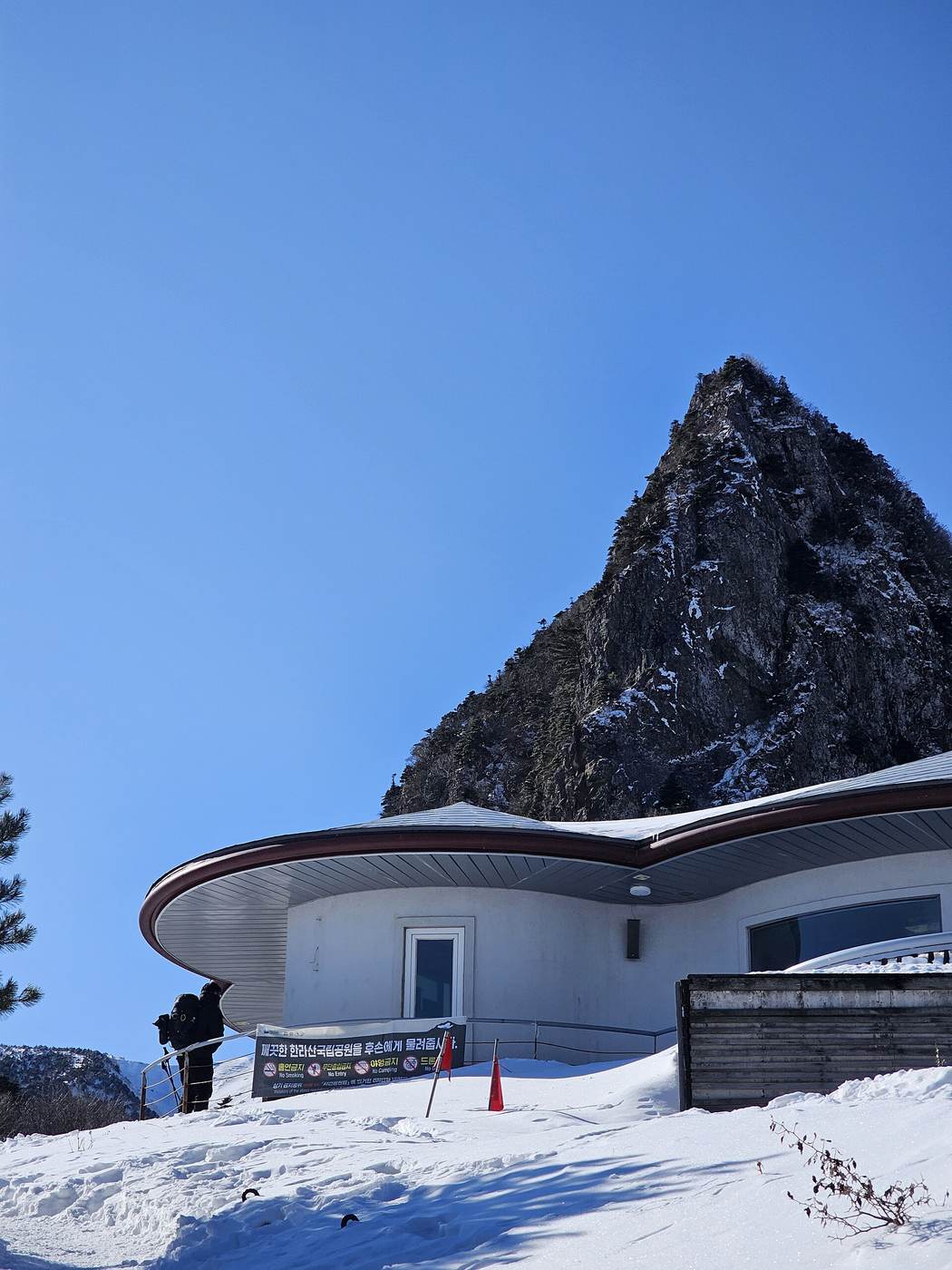 The round, white Samgakbong shelter half buried in snow, with the sharp triangular rock of Samgakbong rising behind it and a hiker with poles by the railing.