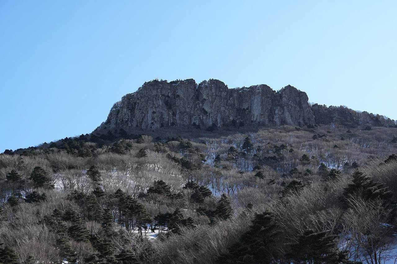 The rock walls of Wanggwanbong on the Gwaneumsa route, dusted with snow, above a slope of bare trees and conifers under a blue sky.