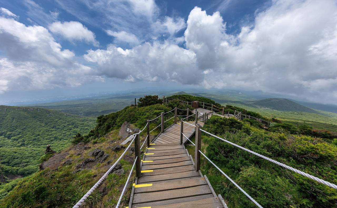 A wooden staircase on the upper Yeongsil trail running through green shrubs, with Jeju's lowlands and oreum under summer clouds beyond.